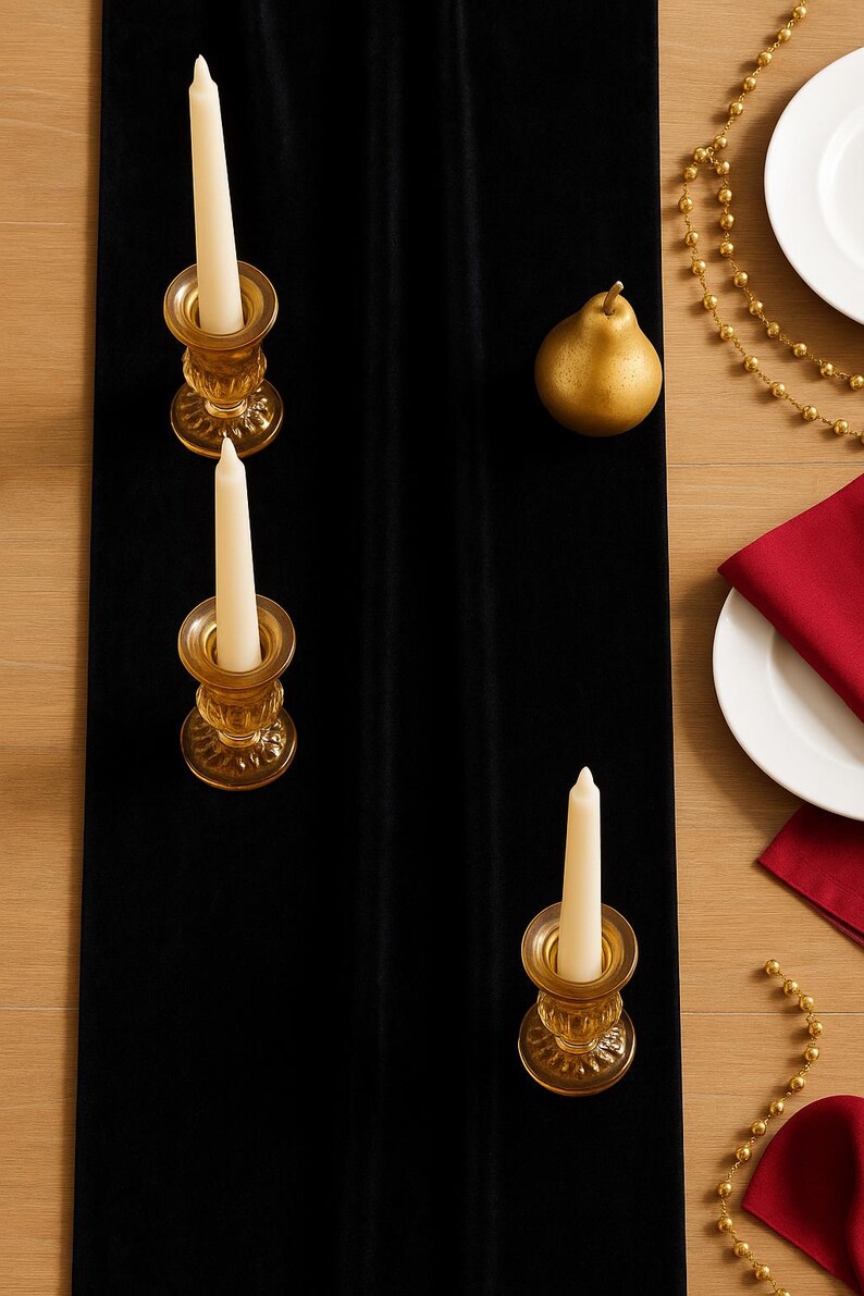 K&ouml;nnte beinhalten: Ein schwarzer Samt-Tischl&auml;ufer mit drei bernsteinfarbenen Glaskerzenhaltern, die jeweils eine wei&szlig;e Kerze halten. Eine goldene Birne und eine goldene Perlenkette liegen auf dem Holztisch. Ein wei&szlig;er Teller und eine rote Serviette sind ebenfalls zu sehen.