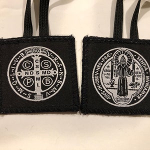 May include: Two black fabric pendants with white embroidered religious symbols and text. Each pendant has a black cord for hanging. One features a cross with the letters C, S, M, and B. The other shows a saint with text.