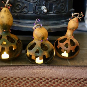 May include: Three gourd jack-o'-lanterns with carved faces and lit candles inside. The gourds are brown and orange, with decorative lights and stems. They sit on a brown rug in front of a dark fireplace, creating a festive Halloween display.