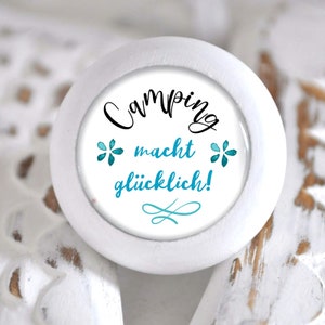 Könnte beinhalten: Weißer runder Holz-Knopf mit einem blau-weißen Aufkleber, auf dem "Camping macht glücklich!" mit einem blauen Schwungmuster steht.