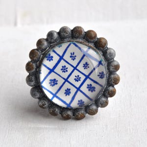 Könnte beinhalten: Runder, dekorativer Schrankknopf mit einem Glasmittelteil, das ein blau-weißes Blumenmuster zeigt. Der Knopf ist von einem Ring aus kleinen, dunkelgrauen und braunen Kugeln umgeben, wodurch ein strukturiertes Design entsteht. Geeignet für Schubladen und Schränke. Durchmesser ca. 4 cm.