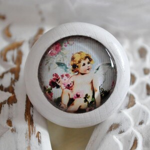 Könnte beinhalten: Weißer Keramik-Knopf mit einem runden, klaren Glaseinsatz, der eine Vintage-Illustration eines geflügelten Cherubins zeigt, der einen Strauß rosa und weißer Blumen hält. Der Cherub ist von einem floralen Muster und einem blau-weiß gestreiften Hintergrund umgeben.