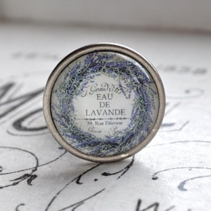 Könnte beinhalten: Ein silberner Knopf mit einer Glasoberfläche, die einen Kranz aus Lavendelblüten und den Text "J. Giraud Fils Eau de Lavande 39, Rue Etienne Grasse Paris" zeigt.
