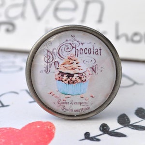 Könnte beinhalten: Ein runder silberfarbener Knopf mit einem Glaseinsatz, der eine Vintage-Illustration eines Schokoladen-Cupcakes mit dem Text "Chocolat" und "Goutez & comparez! Qualite sans Rivale" zeigt.