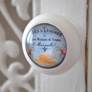 Könnte beinhalten: Weißer Keramik-Knopf mit einem runden, blau-weißen Aufkleber mit einem Strudel aus gelbem Gebäck und dem Text "FES & LEGUMES Rue Mission de France  Moursilley".