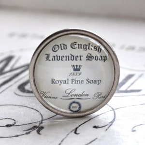 Könnte beinhalten: Ein silberner Knopf mit einer Glasoberfläche, die ein Vintage-Etikett mit der Aufschrift "Old English Lavender Soap 1889 Royal Fine Soap Vienna London Paris 1889" trägt.