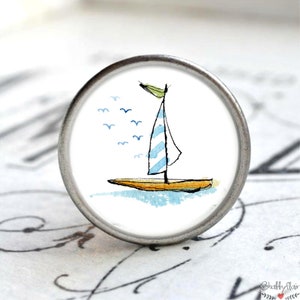Könnte beinhalten: Ein weißer Keramik-Knopf mit einem silbernen Metallring. Der Knopf zeigt eine Aquarell-Illustration eines Segelboots mit einem blau-weiß gestreiften Segel und einem gelben Rumpf. Das Segelboot ist von einer Schar blauer Vögel umgeben.