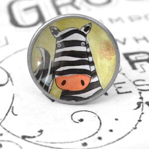 Könnte beinhalten: Ein runder, silberfarbener Knopf mit einer bunten Illustration eines Zebras. Das Zebra hat schwarz-weiße Streifen und eine leuchtend orangefarbene Nase. Der Hintergrund ist hellgelb mit einem dezenten, verblassten Muster.