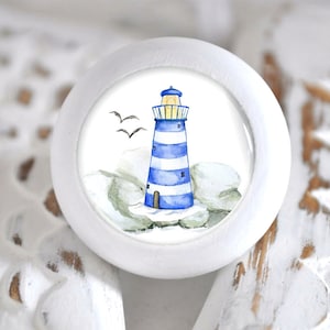 Könnte beinhalten: Weißer Keramik-Knopf mit einer Illustration eines blau-weiß gestreiften Leuchtturms. Der Leuchtturm hat ein gelbes Fenster und ist von weißen Wolken und grauen Felsen umgeben. Zwei weiße Vögel fliegen über dem Leuchtturm.