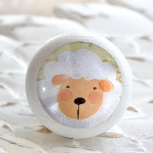 Könnte beinhalten: Weißer Holz-Knopf mit einem runden, bunten Aufkleber eines Cartoon-Schafs mit weißem Gesicht, brauner Nase und rosa Wangen.