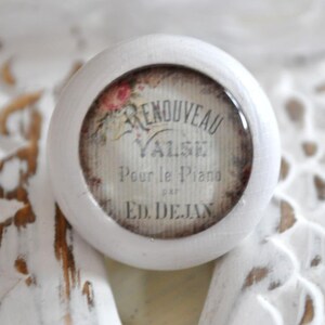 Könnte beinhalten: Ein weißer, runder Schrankknopf mit Vintage-Design. Die Mitte zeigt ein verblasstes Bild mit dem Text "Renouveau Valse Pour le Piano par Ed. Dejan" und floralen Akzenten. Der Knopf ist auf einer strukturierten, weißen Oberfläche platziert.