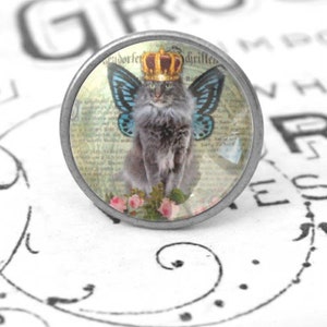 Könnte beinhalten: Ein runder silberfarbener Knopf mit einer Glasoberfläche, die eine graue Katze mit einer Krone und blauen Schmetterlingsflügeln zeigt. Die Katze sitzt auf einem floralen Hintergrund mit einem Vintage-Zeitungsdruck.