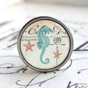 Könnte beinhalten: Ein silberner Knopf mit einer Glasoberfläche, die eine Vintage-Illustration eines blauen Seepferdchens und zweier rosa Seesterne zeigt. Das Bild befindet sich auf einem cremefarbenen Hintergrund mit dem sichtbaren Text "Correspondance" und "Modèle".