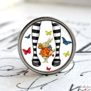Könnte beinhalten: Ein weißer Keramik-Knopf mit einem schwarz-weißen Bild von Alice im Wunderland's Beinen und dem weißen Kaninchen. Das Bild enthält auch Schmetterlinge in verschiedenen Farben.