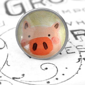 Könnte beinhalten: Runder, silberfarbener Schrankknopf mit einer Aquarellillustration eines Schweinegesichts. Das Schwein hat eine rosa Schnauze, rosige Wangen und kleine schwarze Augen. Der Hintergrund ist hellgelb.
