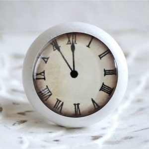 Könnte beinhalten: Weißer runder Knopf mit einem Vintage-Zifferblatt mit römischen Ziffern. Das Zifferblatt hat einen cremefarbenen Hintergrund mit schwarzen Zeigern und schwarzen römischen Ziffern.