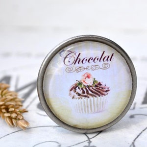 Könnte beinhalten: Ein silberner Knopf mit einem runden Glaseinsatz, der eine Vintage-Illustration eines Schokoladen-Cupcakes mit einer rosa Rose oben zeigt. Der Text "Chocolat" ist in einer dekorativen Schrift über dem Cupcake geschrieben.