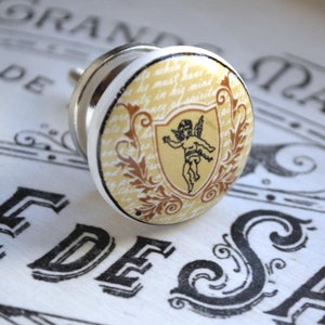 Könnte beinhalten: Ein weißer Keramik-Knopf mit einem goldenen und braunen Cherub-Design. Der Cherub hält einen Schild mit einem floralen Design. Der Knopf hat eine silberne Schraube.