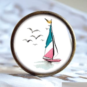 Könnte beinhalten: Ein weißer Keramik-Knopf mit einem bronzefarbenen Metallrand zeigt eine Aquarell-Illustration eines Segelboots mit einem rosa und türkisfarbenen Segel und einer gelben Flagge. Der Hintergrund enthält eine Aquarell-Waschung in Blau und Grün sowie eine Schar schwarzer Vögel, die fliegen.