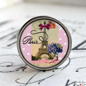 Könnte beinhalten: Ein silberner Knopf mit rosa und weißem Hintergrund, der ein Vintage-Postkartenbild des Eiffelturms in Paris, Frankreich, zeigt. Die Postkarte hat eine rosa Schleife und das Wort "Paris" ist in Kursivschrift geschrieben.