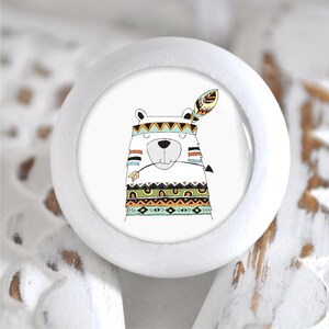 Könnte beinhalten: Weißer Keramik-Knopf mit einer bunten Illustration eines Bären, der ein Kopfschmuck mit einer Feder trägt. Der Bär trägt eine bunte gemusterte Weste.