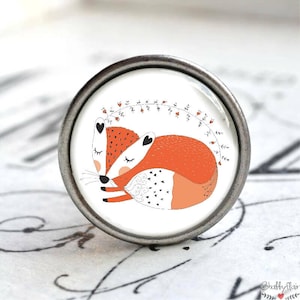 Könnte beinhalten: Runder, silberfarbener Schubladengriff mit einer Illustration von zwei schlafenden Füchsen. Die orange-weißen Füchse mit schwarzen Herzakzenten liegen auf weißem Grund. Ein Blumengirlandebogen befindet sich über ihnen.