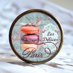 Könnte beinhalten: Ein runder, bronzefarbener Knopf mit einem Glaseinsatz, der einen Stapel von drei Macarons in rosa, lila und beige zeigt. Das Bild enthält auch ein Etikett im Vintage-Stil mit dem Text "Les Délices Paris" auf Französisch.