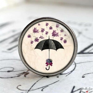 Könnte beinhalten: Ein runder, silberfarbener Knopf mit einem Vintage-Bild eines schwarzen Regenschirms, aus dem violette Blumen fallen. Der Regenschirm hat einen Strauß violetter Blumen am Griff.