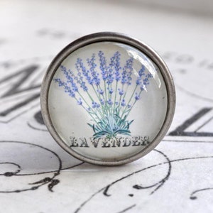 Könnte beinhalten: Ein silberner Knopf mit einer Glasoberfläche, die eine Illustration einer Lavendelblüte und den darunter gedruckten Schriftzug "LAVENDER" zeigt.