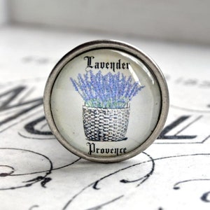 Könnte beinhalten: Ein silberner Knopf mit einem Glaseinsatz, der eine lilafarbene Lavendelpflanze in einem geflochtenen Korb zeigt. Der Text "Lavender Provence" ist unter dem Bild gedruckt.
