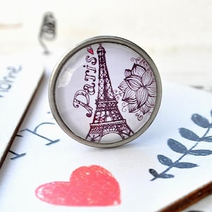Könnte beinhalten: Ein silberner Ring mit einem Glascabochon, der eine Schwarzweißillustration des Eiffelturms in Paris, Frankreich, zeigt. Das Wort "Paris" ist in Kursivschrift auf dem Cabochon geschrieben. Eine rosa Blume ist ebenfalls auf dem Cabochon abgebildet.