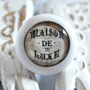Könnte beinhalten: Weißer runder Knopf mit einem Glaseinsatz mit schwarzem Text, der "Maison de Luxe 19, Rue de Fontaine PARIS" lautet.