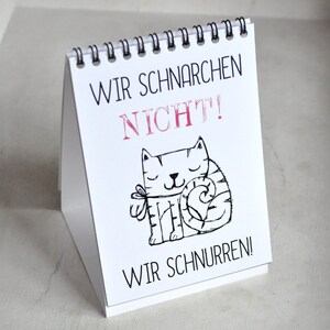May include: A white desk calendar with a black spiral binding. The calendar features a black and white illustration of a cat with the text "Wir schnarchen nicht! Wir schnurren!" in German.