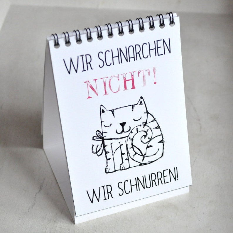May include: A white desk calendar with a black spiral binding. The calendar features a black and white illustration of a cat with the text "Wir schnarchen nicht! Wir schnurren!" in German.