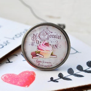 Könnte beinhalten: Ein runder silberfarbener Anhänger mit einem Vintage-Bild eines rosa und weißen Cupcakes mit Schokoladenstreuseln. Der Text "Chocolat Poulain" in Französisch ist sichtbar, sowie der Spruch "Goutez & comparez Qualite sans Rivale".