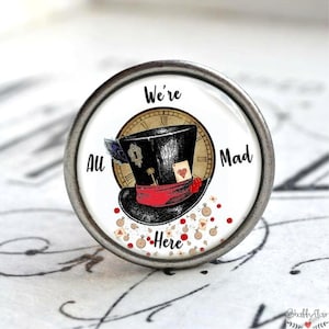 Könnte beinhalten: Ein weißer runder Knopf mit einem silbernen Metallrand zeigt eine Schwarzweißillustration des Hutmachers mit einem roten Band und einem Zifferblatt. Der Text "We're All Mad Here" ist um den Hut herum gedruckt.