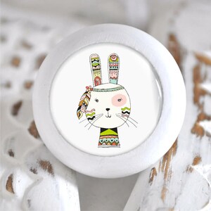 Könnte beinhalten: Weißer Keramik-Knopf mit einer bunten Illustration eines Kaninchens, das ein Stirnband mit Federn und geometrischen Mustern trägt.
