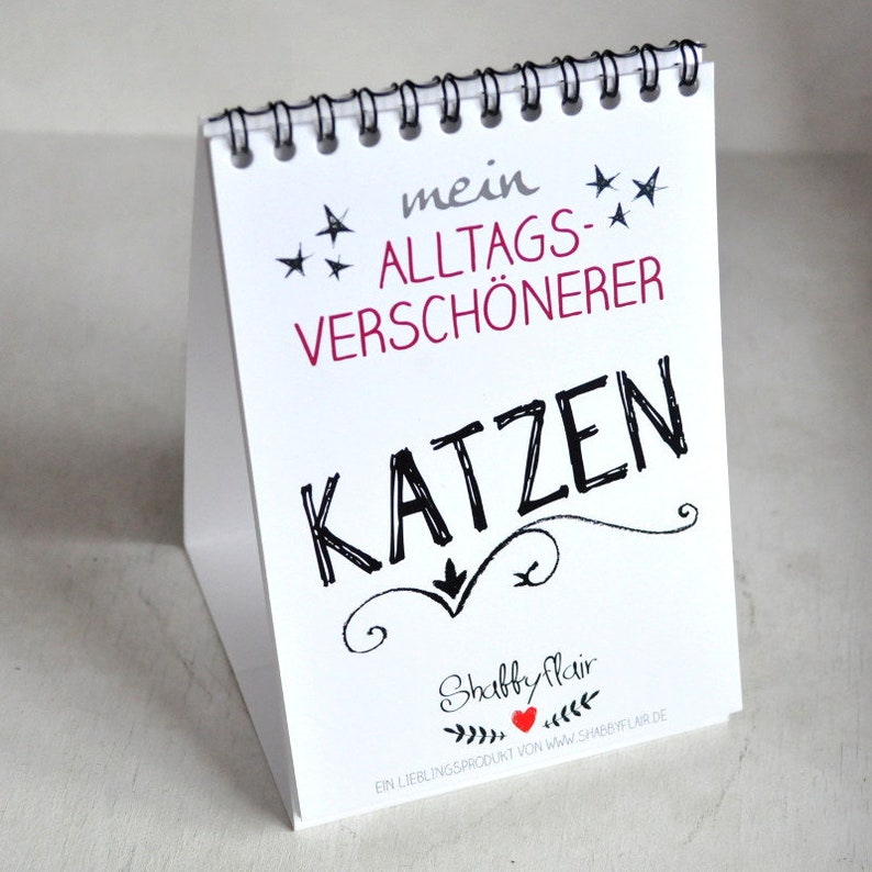 May include: White tabletop easel with a black spiral binding. The easel has a black and white graphic of a cat with the text "mein ALLTAGS-VERSCH&Ouml;NERER KATZEN" and the text "Shabby Flair EIN LIEBLINGSPRODUKT VON www.shabbyflair.de".