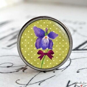 Könnte beinhalten: Eine violette Blume mit einem grünen Stiel und einer roten Schleife auf grünem Hintergrund mit weißen Punkten. Die Blume befindet sich in einem silbernen Metallrahmen.
