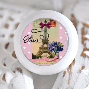 Könnte beinhalten: Weißer runder Knopf mit rosa und weißem Hintergrund, der ein Vintage-Postkartenbild des Eiffelturms in Paris, Frankreich, zeigt. Die Postkarte hat ein rosa Band und das Wort "Paris" in Kursivschrift.