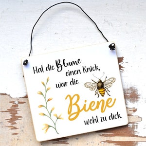 May include: A white wooden sign with a black string for hanging. The sign has a yellow flower and a bee illustration. The text on the sign reads "Hat die Blume einen Knick, war die Biene wohl zu dick."