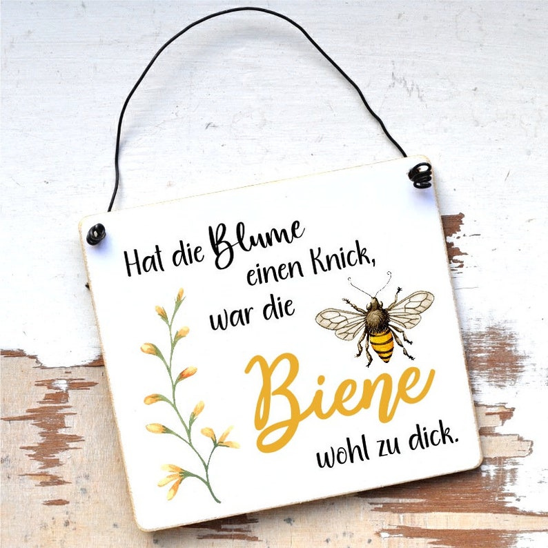 May include: A white wooden sign with a black string for hanging. The sign has a yellow flower and a bee illustration. The text on the sign reads "Hat die Blume einen Knick, war die Biene wohl zu dick."