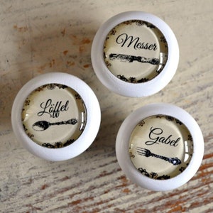 Könnte beinhalten: Drei weiße runde Knöpfe mit schwarz-weißen Bildern von Besteck und den Worten "Messer", "Löffel" und "Gabel" darauf.