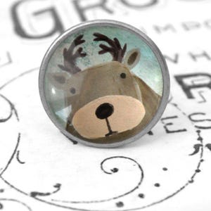 Könnte beinhalten: Runder, silberfarbener Schrankknopf mit einem Cartoon-Rentiergesicht. Das Rentier hat braune Geweihe, eine beige Schnauze und eine schwarze Nase. Der Hintergrund ist hellblau mit Aquarell-Effekt.