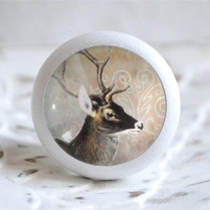 Könnte beinhalten: Weißer, runder Schrankknopf mit einer Vintage-Illustration eines Hirsches. Der Hirsch, in Braun und Schwarz, hat ein großes Geweih und ist vor einem beigen, gemusterten Hintergrund platziert. Der Knopf ist aus Holz.