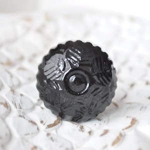Könnte beinhalten: Schwarzer Glas-Schrankknopf mit geometrischem Muster. Der Knopf hat eine erhabene Mitte und einen gewellten Rand.