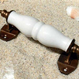 Könnte beinhalten: Weißer Keramik-Türgriff mit Messing-Akzenten. Der Griff ist oval geformt und hat eine glatte, polierte Oberfläche. Die Messing-Akzente befinden sich an den Enden des Griffs und sind ebenfalls glatt und poliert.