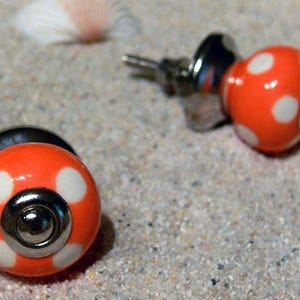 Könnte beinhalten: Nahaufnahme von zwei orangefarbenen Ohrsteckern mit weißen Punkten. Die Ohrringe haben einen silbernen Stecker und Verschluss. Die Ohrringe liegen auf einer sandigen Oberfläche, mit einer Muschel im Hintergrund.