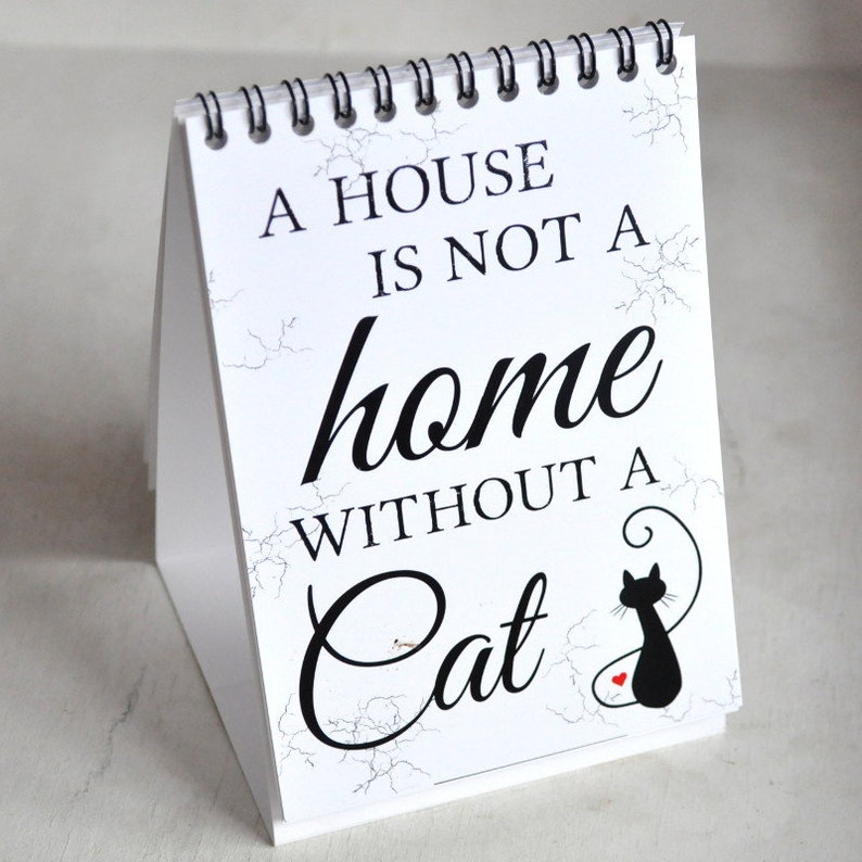 May include: A white desk calendar with a black and white marble pattern. The text on the calendar reads "A house is not a home without a cat." A black silhouette of a cat with a red heart is in the bottom right corner.