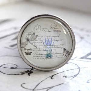 Könnte beinhalten: Ein silberner Knopf mit einer Glasoberfläche, die mit einem Vintage-Blumendesign mit Lavendelblüten und einer Libelle verziert ist. Die Glasoberfläche trägt eine handschriftliche Inschrift mit der Aufschrift "Dragonfly" und "Maker".